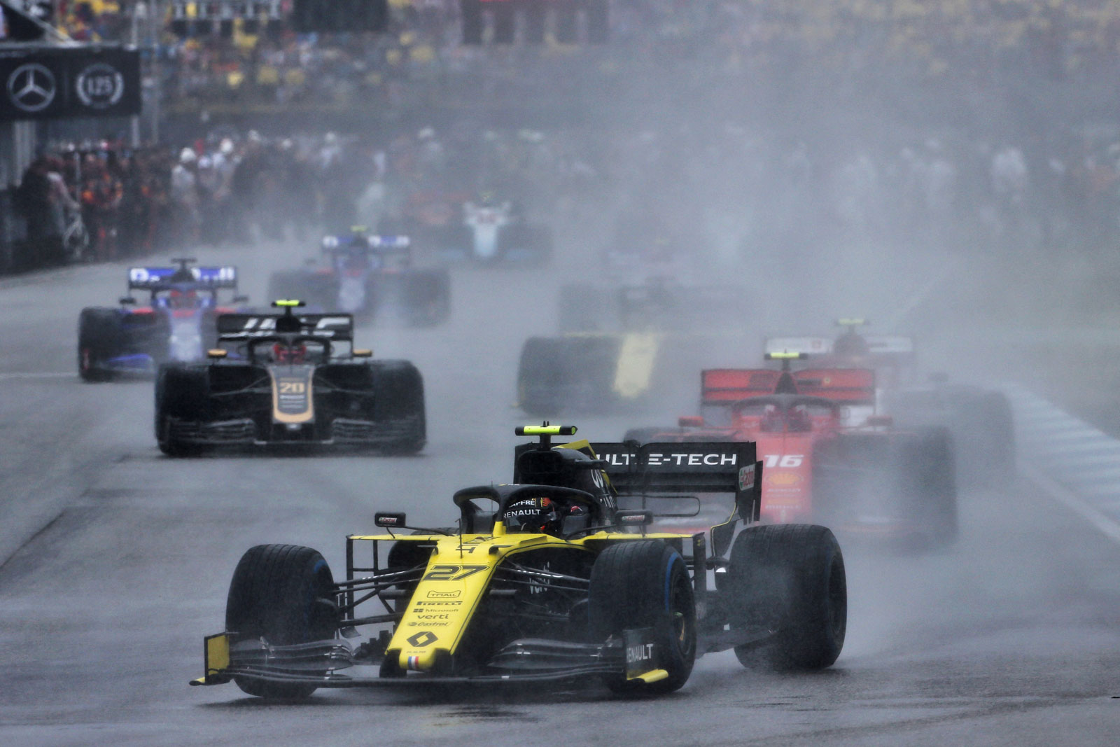 A yellow Renault car in the foreground leads multiple other Formula 1 cars in a rainy race
